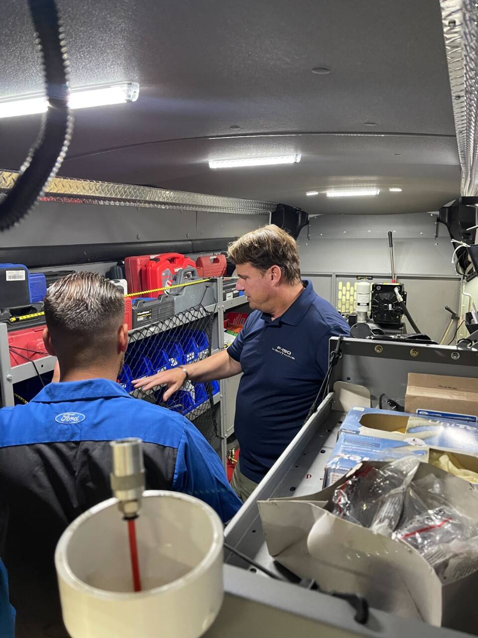 Two men inside a van look at vehicle parts.